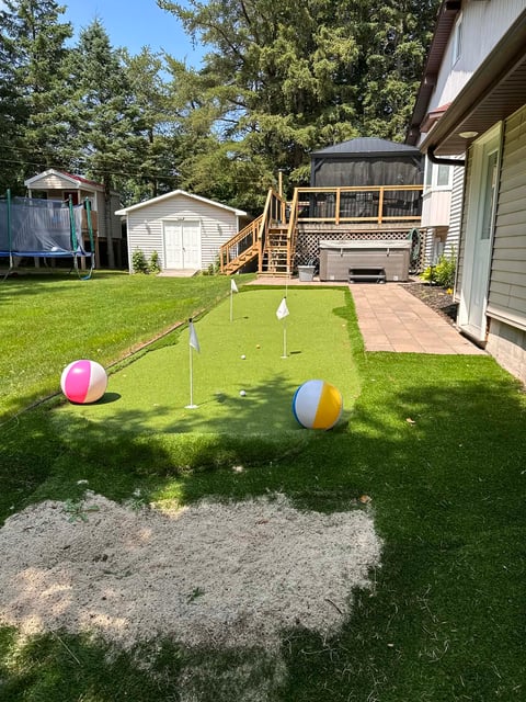 Residential backyard with a mini golf course setup on grass, featuring colored balls and flagged holes, with a deck and shed in the background