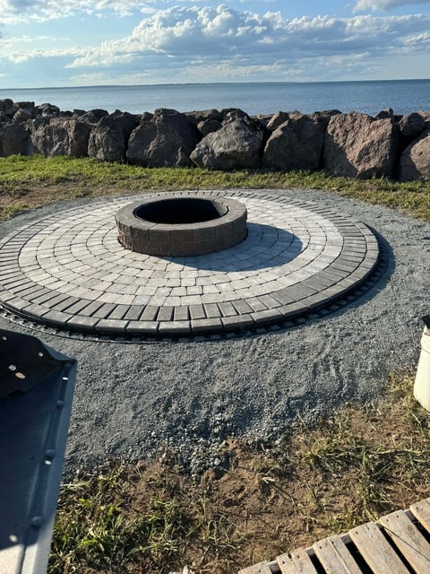 Circular stone fire pit with patterned pavers on a beach overlooking rocky shores and water under cloudy sky