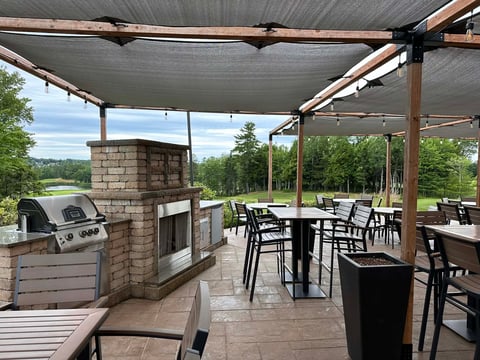 Covered outdoor patio with dining tables, chairs, brick grill station, and scenic tree and lake views