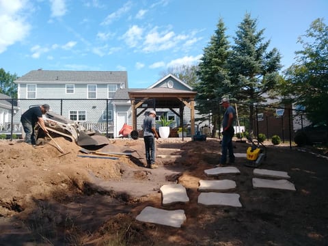 Construction workers installing pavers in a residential backyard with houses and trees in background