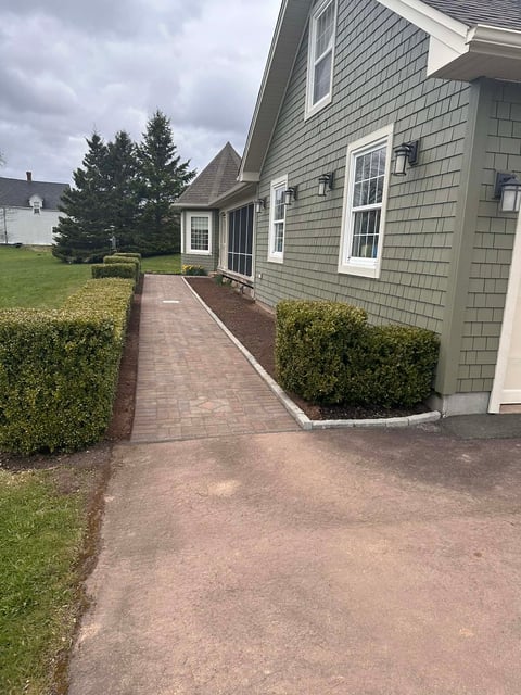 Gray shingled house with brick walkway, manicured shrubs, and green lawn under cloudy sky