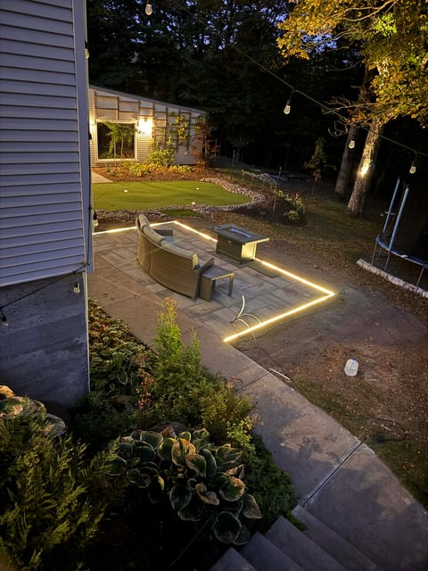 Nighttime backyard patio with LED-lit pathway, built-in seating, lush landscaping, and illuminated house in background