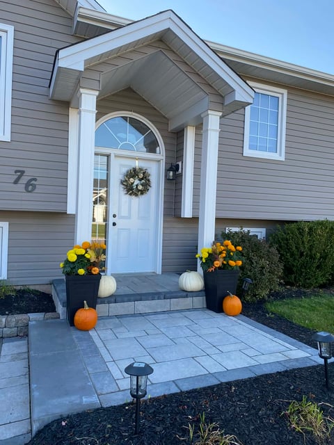 Front entrance of a house with white door, gray siding, and decorative fall pumpkins and yellow flowers flanking the paved walkway.