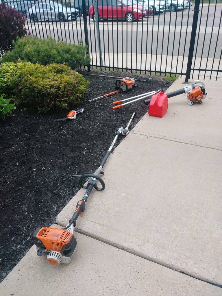 Outdoor yard work equipment arranged on concrete and mulch, including string trimmer, hedge trimmer, gas can, and power tools
