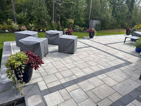 Modern patio with geometric paving, gray furniture covers, potted plants, and wooded background